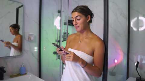 Video of a brunette woman in a white towel, holding a phone, standing in a modern, white-tiled bathroom with a mirror and fluorescent lights.