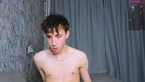 Video of a shirtless young man with short, tousled brown hair and fair skin, wearing a silver necklace, looking introspective. Background features gray curtains and a TV screen displaying colorful text.