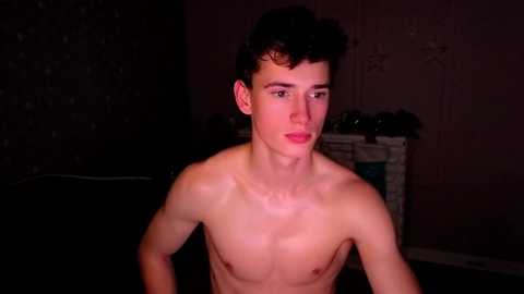 Video of a shirtless young white male with curly dark hair, standing indoors with dim lighting, looking to the left, showing a muscular physique and pale skin. Background features a dark, textured wall with decorative star patterns.