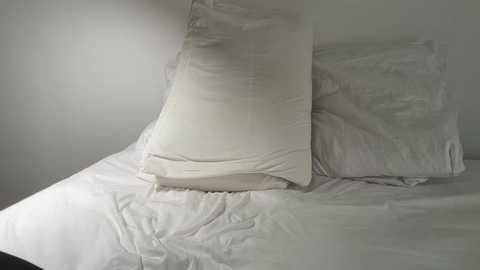A video of a neatly made bed with white linens and a single white pillow on a gray wall. The bed is in a minimalist room, with soft, diffused lighting creating a serene atmosphere.