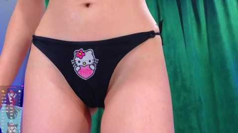 Video of a light-skinned woman's lower torso, wearing black panties with a pink heart and white Hello Kitty design, standing against a green curtain background.