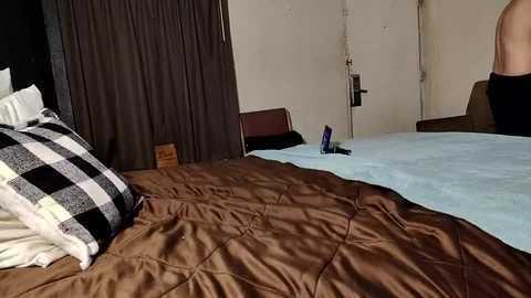 Video of a bedroom with a neatly made bed covered in a brown and white checkered quilt. The room has a beige wall and a dark curtain in the background.
