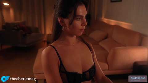 Media: Video of a brunette woman in black lingerie, standing in a dimly lit, warmly-lit living room with beige couch and lamp.