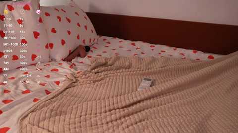 Media: A video of a bedroom with a beige knitted blanket over a bed adorned with white sheets featuring red heart patterns. A wooden headboard and a white pillow with the same heart pattern are visible.
