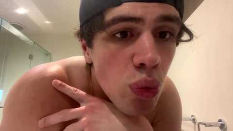 Video of a shirtless young man with light skin and short dark hair, wearing a backwards baseball cap, puckering his lips, and flashing a peace sign. Background shows a modern bathroom with a mirror and sink.