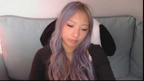 Media: Video of a young Asian woman with long, wavy, lavender hair, wearing a black top, sitting on a light blue sofa.