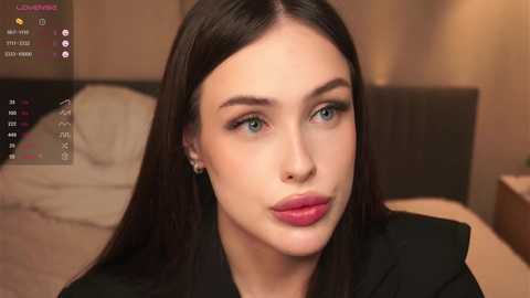 A video of a young woman with fair skin, long dark hair, and striking blue eyes, wearing a black top, in a bedroom with a bed and warm lighting.