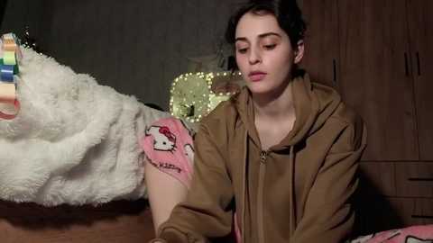 Video of a young woman with short, curly black hair, wearing a brown hoodie with a pink Hello Kitty sleeve, sitting on a wooden floor beside a plush white dog.