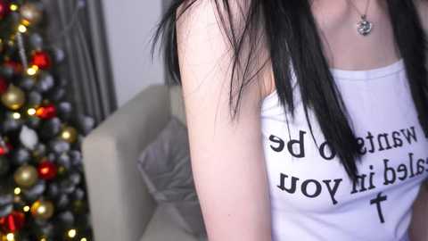 Video of a young woman with pale skin, long black hair, wearing a white tank top with Spanish text, standing in a room with a Christmas tree adorned with red and gold ornaments.