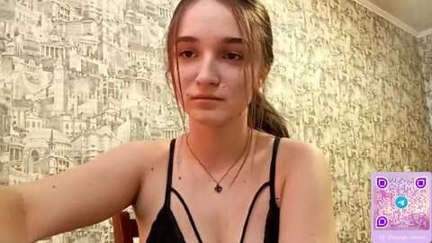 Video of a young Caucasian woman with light skin, brown hair tied back, wearing a black strappy top, sitting in a room with a textured wallpaper pattern. She has a neutral expression.