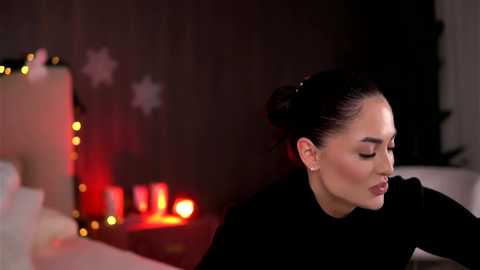 Video of a woman with dark hair in a bun, wearing a black sweater, looking thoughtful in a dimly lit room with red and white holiday decorations.