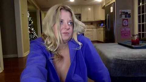 Video of a blonde woman with wavy hair, wearing a low-cut blue shirt, sitting in a cozy living room with a Christmas tree and kitchen in the background.