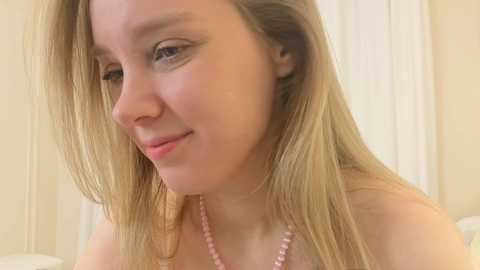Video of a young, fair-skinned woman with long, straight blonde hair, wearing a pink beaded necklace, smiling softly, in a softly lit, beige room.