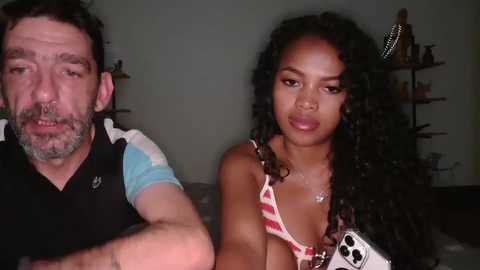Video of a Caucasian man with a beard and glasses, and a Black woman with curly hair, both sitting on a bed in a dimly lit room.