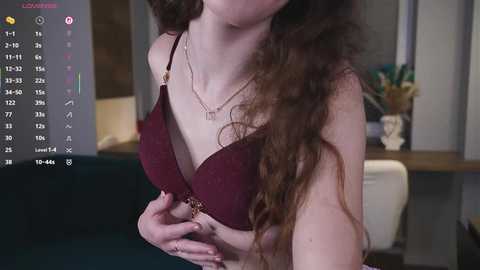 Video of a fair-skinned woman with long, curly brown hair, wearing a burgundy lace bra, standing indoors near a desk with a vase.