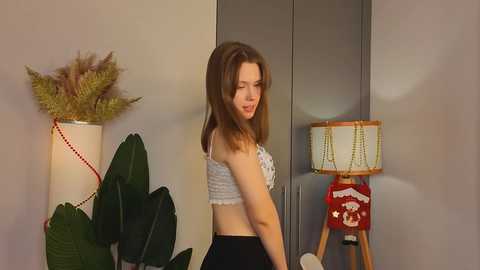 Media: Video of a young woman with straight brown hair, wearing a white lace bralette and black pants, standing in a modern room with a grey wall, fern, and lamp.