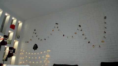 Video of a modern, minimalist room with a white brick wall adorned with small, hanging artwork and string lights. A red hat and black backpack hang on a white shelf.