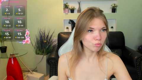 Video of a young Caucasian woman with light skin, long blonde hair, wearing a white lace dress, sitting in a black leather chair. Background includes green walls, potted plants, and a digital temperature display.