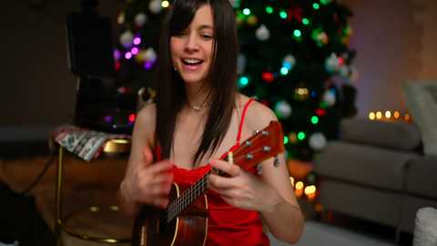 Video of a joyful woman with long brown hair, playing a ukulele in a festive living room with a Christmas tree and colorful lights in the background.