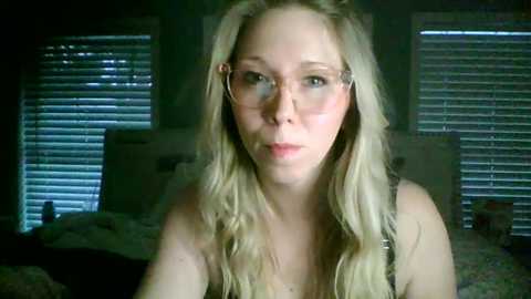 Video of a blonde, fair-skinned woman with long wavy hair, wearing glasses, in a dimly lit bedroom with closed window blinds, and a messy bed.