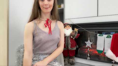 Video of a smiling young woman with light skin and long brown hair, wearing a gray tank top and silver tinsel skirt, standing in front of a Christmas-themed fireplace with a Santa doll and festive decorations.