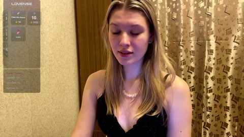 Media: Video of a young, fair-skinned woman with long, straight blonde hair, wearing a black lace bra, standing in a bathroom with beige wallpaper and patterned shower curtain.