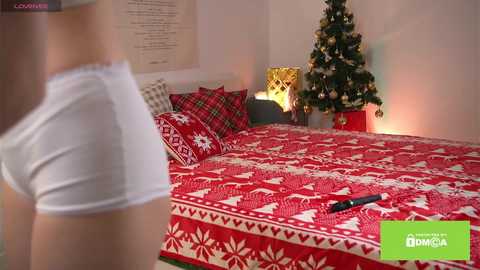 A video of a cozy, festive bedroom with a Christmas tree, red-patterned bedding, and a woman in white underwear on the left side.