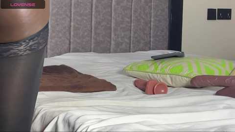 Video of a bedroom with a gray cushioned headboard, a white bed, and a person wearing gray lace lingerie, with a pink dildo and green pillow nearby.