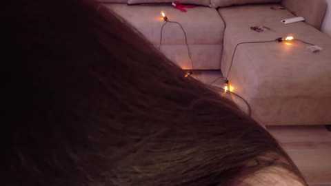 Video of a long, dark brown hair lying on a beige sofa, adorned with string lights, remote control, and red object near the cushion.