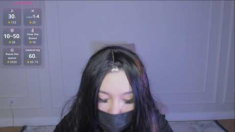 Video of a young woman with long black hair, wearing a black face mask, standing in a dimly lit room with white walls and a display showing vital signs.