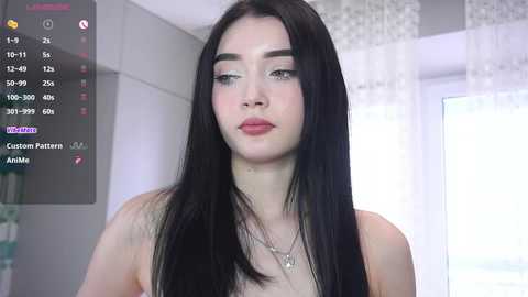Video of a young woman with long black hair and fair skin, wearing a necklace, in a light-colored room.