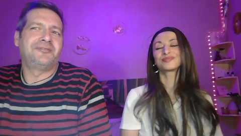 Video of a smiling middle-aged man with short hair in a striped shirt, and a brunette woman with long hair, both in a dimly-lit, cozy room with purple lighting, shelves, and a bed.