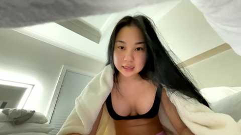 Video of an East Asian woman with long black hair, wearing a black bra and white robe, lying on a bed in a modern bedroom with white walls and minimalistic decor.