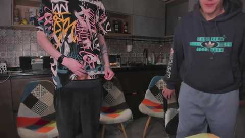 Video of a man with a large, erect penis in a colorful, patterned T-shirt, standing in a modern kitchen with tiled backsplash and chairs.