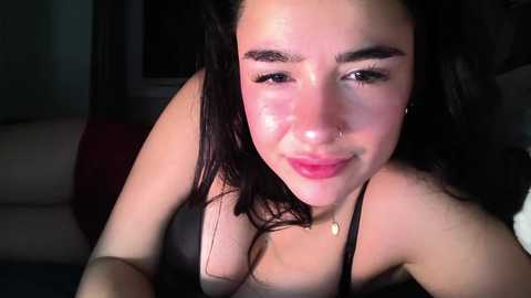 Video of a young woman with fair skin, long dark hair, and a nose ring, smiling, wearing a black bra, indoors with dim lighting.