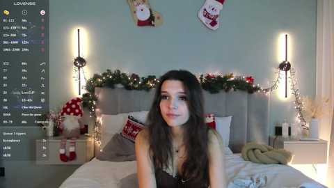 A video of a young woman with long dark hair, wearing a black lace top, sitting on a bed with festive decorations. The room is warmly lit with string lights and Christmas ornaments.