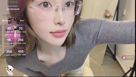 Video of a young, fair-skinned woman with shoulder-length brown hair, wearing clear glasses and a grey ribbed sweater, taken in a modern, well-lit room with a video game interface overlay.