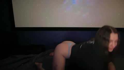 A video of a woman with long brown hair, wearing a black leather outfit, on all fours on a bed, with a dimly lit room and a projector screen displaying a starry sky in the background.