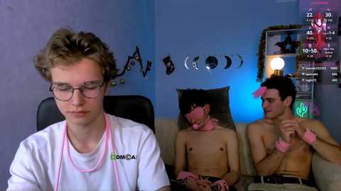 Video of two shirtless teenage boys in a dimly lit room, one wearing pink hair scrunchies and a white t-shirt, the other with a serious expression, both sitting on a couch.