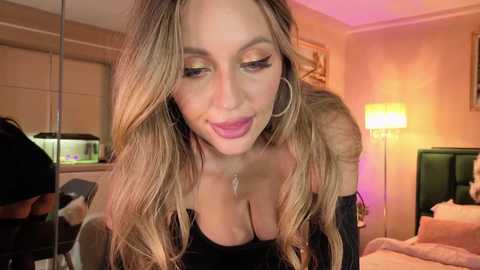 Video of a smiling woman with long, wavy blonde hair, wearing a black dress with a deep neckline, sitting in a warmly lit bedroom with a green bed, white lamp, and aquarium.