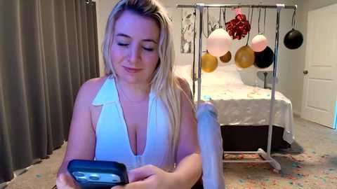 Video of a blonde woman in a white halter top, smiling, texting on a black phone in a hospital room with a bed, balloons, and festive decorations.