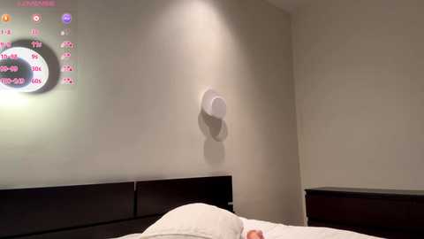 Video of a modern, minimalist bedroom with beige walls, a dark wooden headboard, and a digital clock mounted on the wall. A round, white light fixture is positioned above the bed, which is neatly made with white linens.