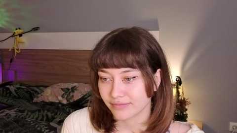 Video of a young woman with shoulder-length brown hair and light skin, smiling slightly, standing in a bedroom with a wooden headboard, green bedspread, and a yellow lamp.