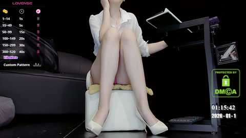 Video of a slender woman with fair skin, wearing a white blouse and white high heels, sitting on a white cushioned chair. She is on a black stage with a black screen and a desk with a laptop.