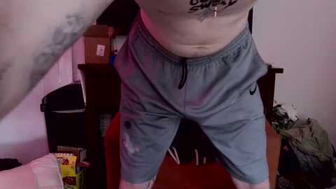 Video of a tattooed, shirtless, fair-skinned person wearing light blue athletic shorts with a Nike logo, standing indoors with a wooden shelf, a red light, and a green object in the background.