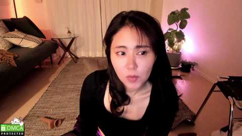 Media: Video of a young Asian woman with long black hair, wearing a black top, seated on a textured rug in a cozy living room with a green plant, dark furniture, and dim lighting.