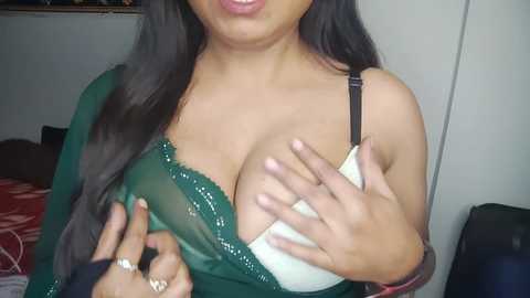 Media: A video shows a woman with long dark hair, medium skin tone, wearing a green, semi-transparent dress, revealing a white bra. Her hands are cupping her breasts, and a ring is visible on her finger. The background is a simple bedroom with a red-patterned blanket.