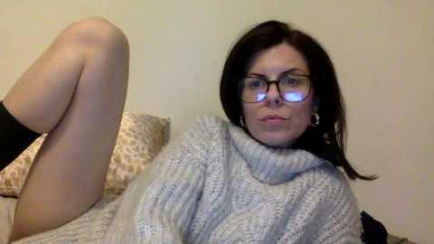 A video of a woman with fair skin, dark hair, and glasses, lounging on a bed in a cozy, light-colored room, wearing a grey, textured sweater and black socks.