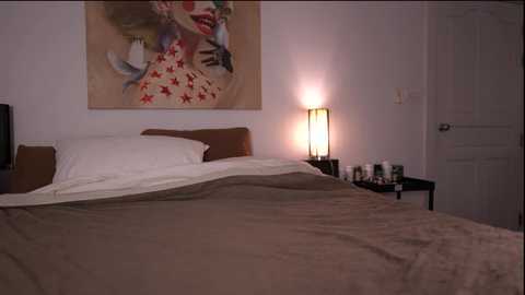 Video of a minimalist bedroom with a beige bedspread, white pillows, a large abstract painting, a black side table with a lamp, and a closed white door.