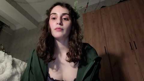 Video of a young woman with curly brown hair, wearing a dark green robe, standing in a dimly lit bedroom with wooden wardrobes and a bed visible in the background.